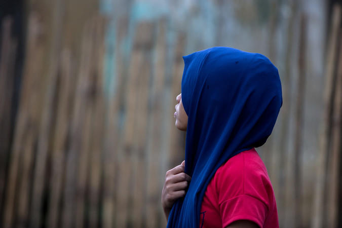 A girl in the woods wearing a blue hijab looks away from the camera.