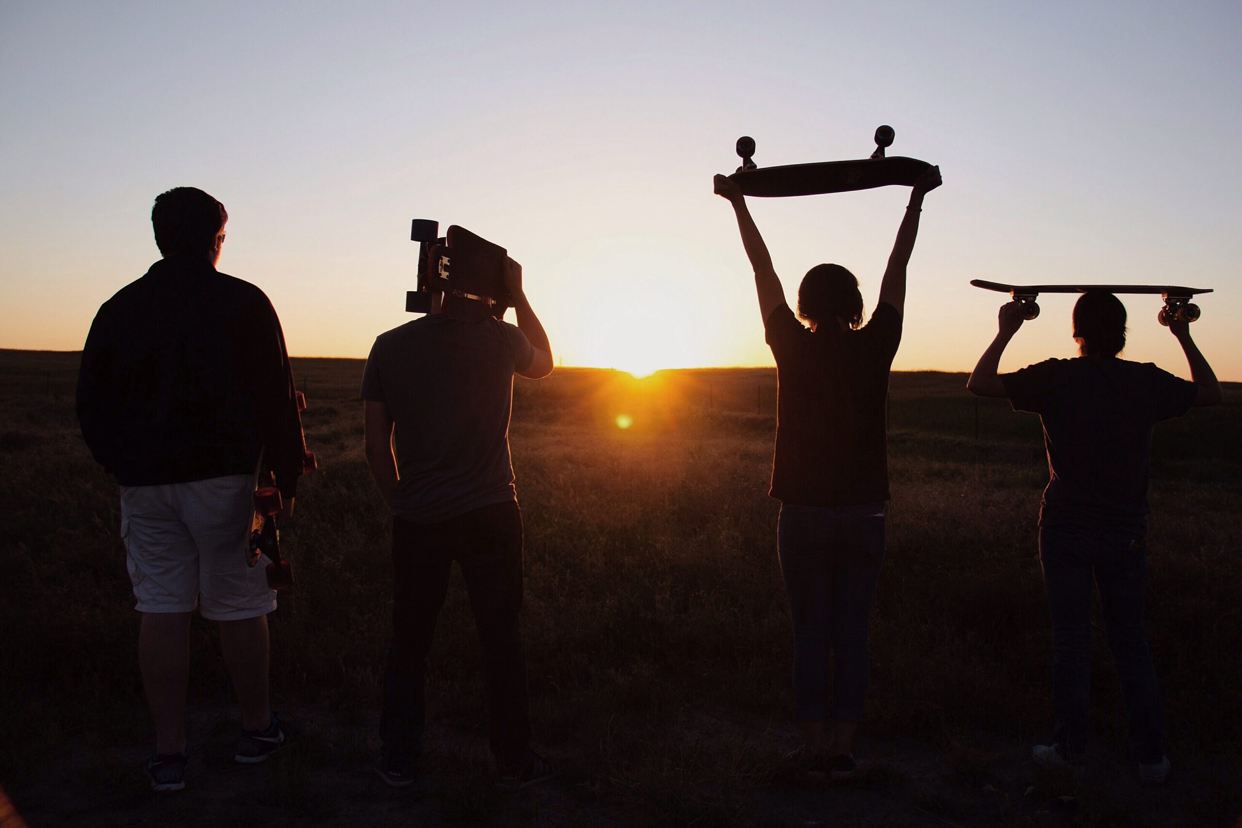 Four young people silhouetted against a sunset, two hold skateboards