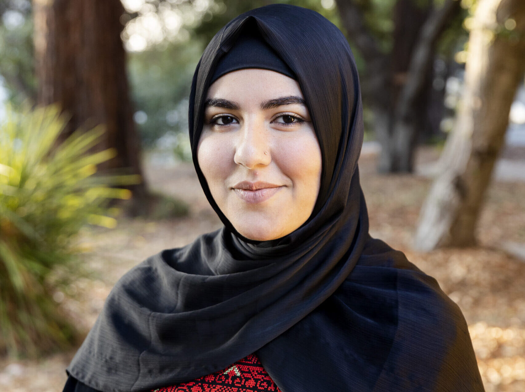 Portrait of a woman in a black hijab with a traditional Palestinian abaya with red stitching.