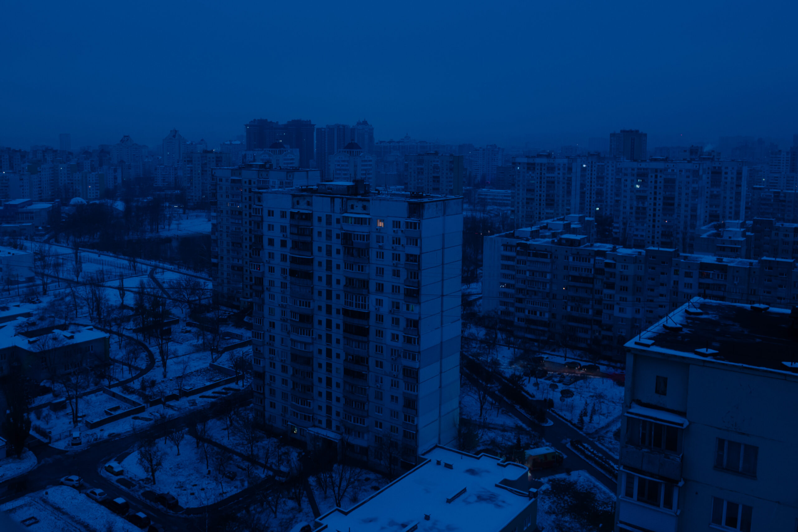 A city in almost total darkness, with ambient light depicting a blue hue. There is snow on the ground.