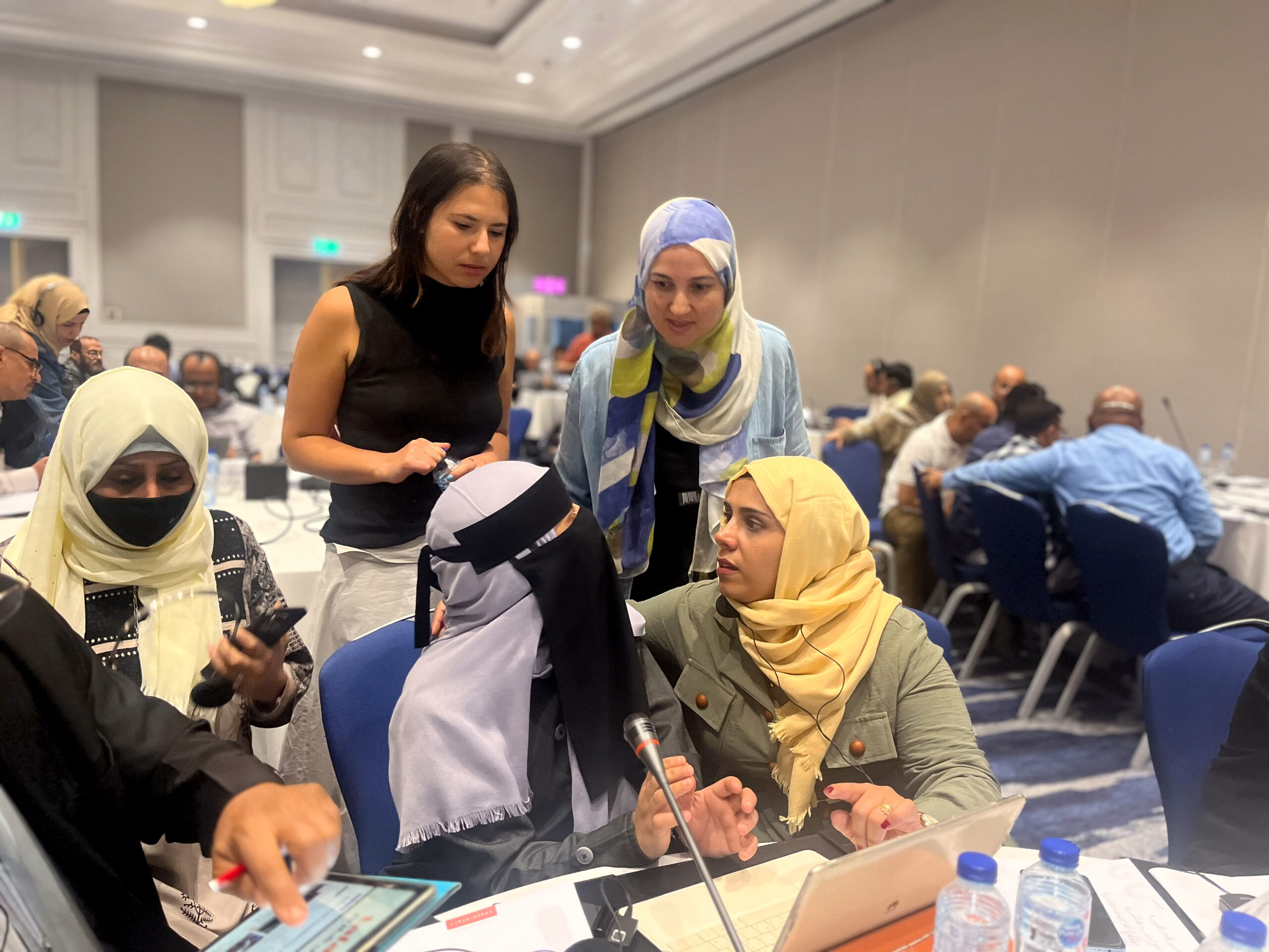 A group of women work together in a conference setting. Most of the women are wearing a hijab, and one woman is not wearing a hijab. She has brown hair.