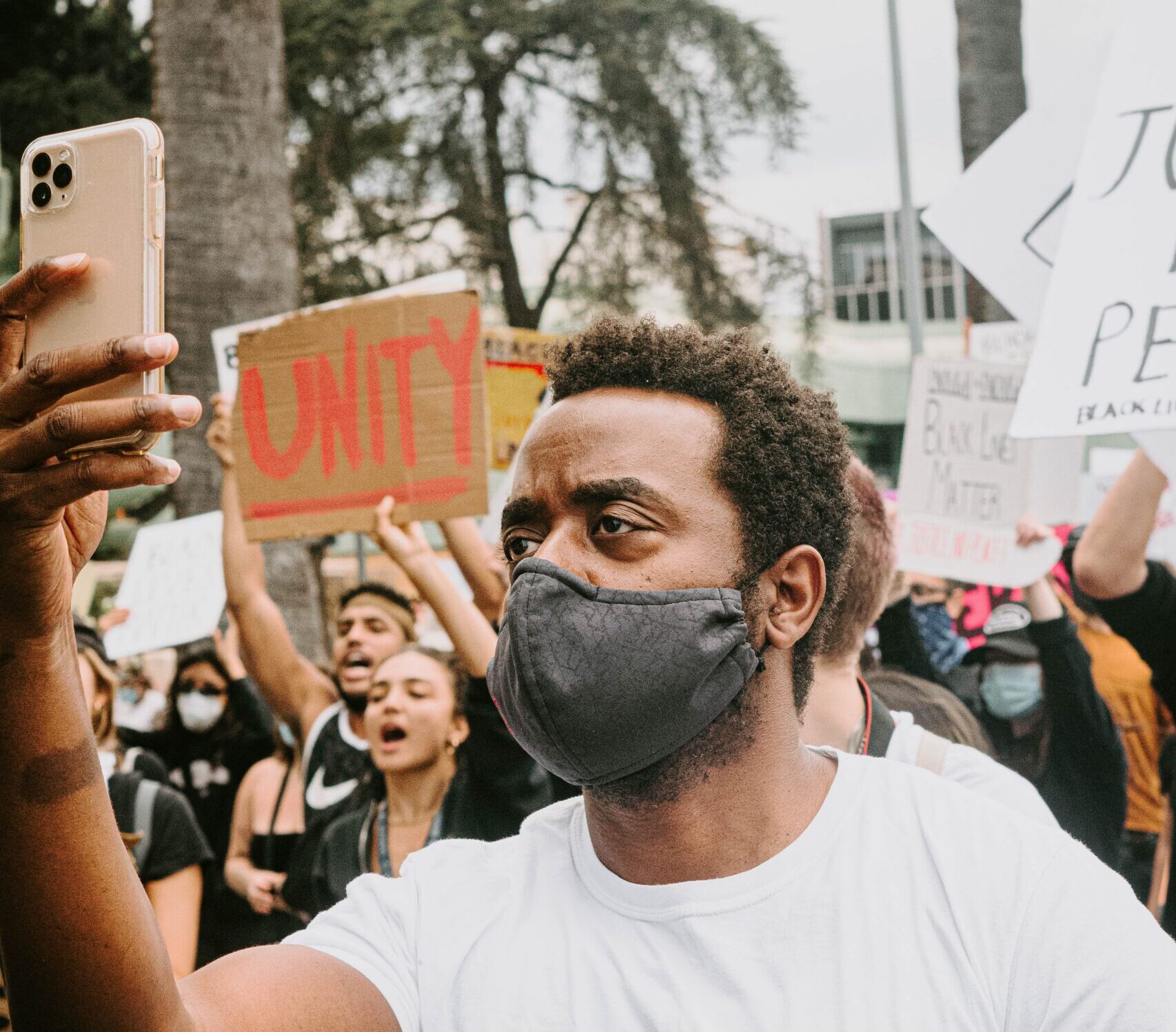 A man with brown hair and a mask uses his phone at a protest, a sign with the word 