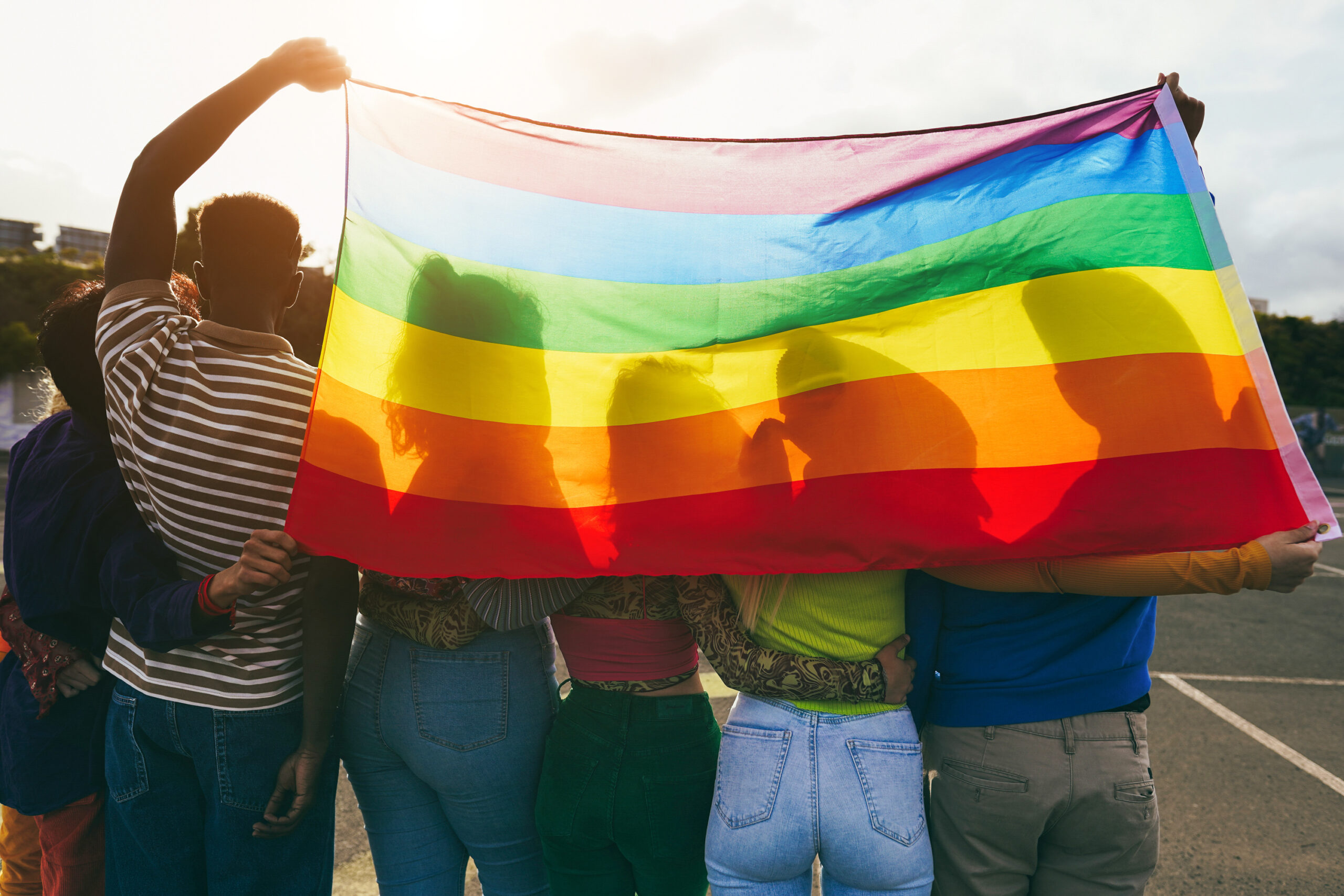 People hold a rainbow flag up to the sunlight, their silhouettes can be seen behind it.