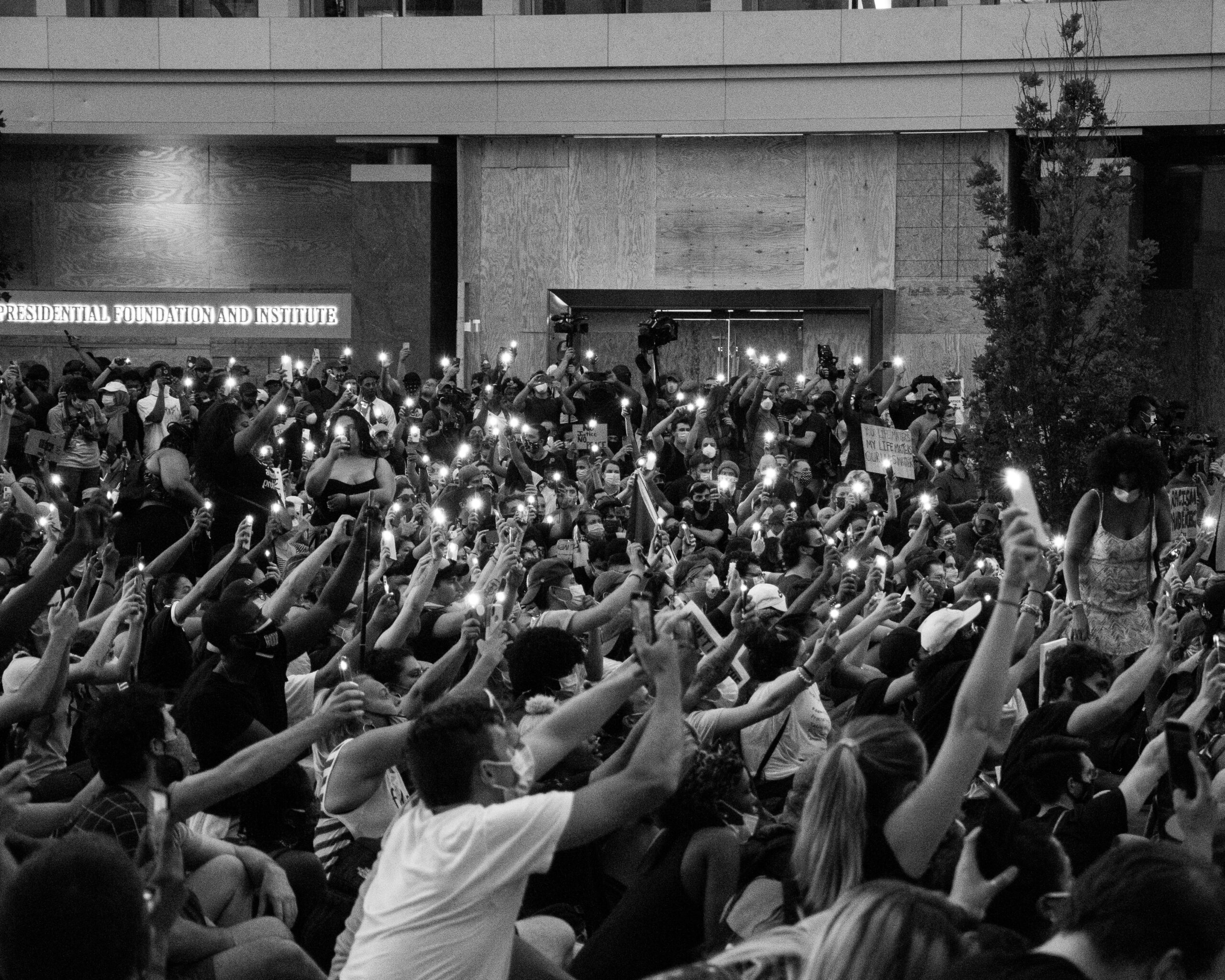A black and white image of protesters wearing masks using their phones as flashlights.