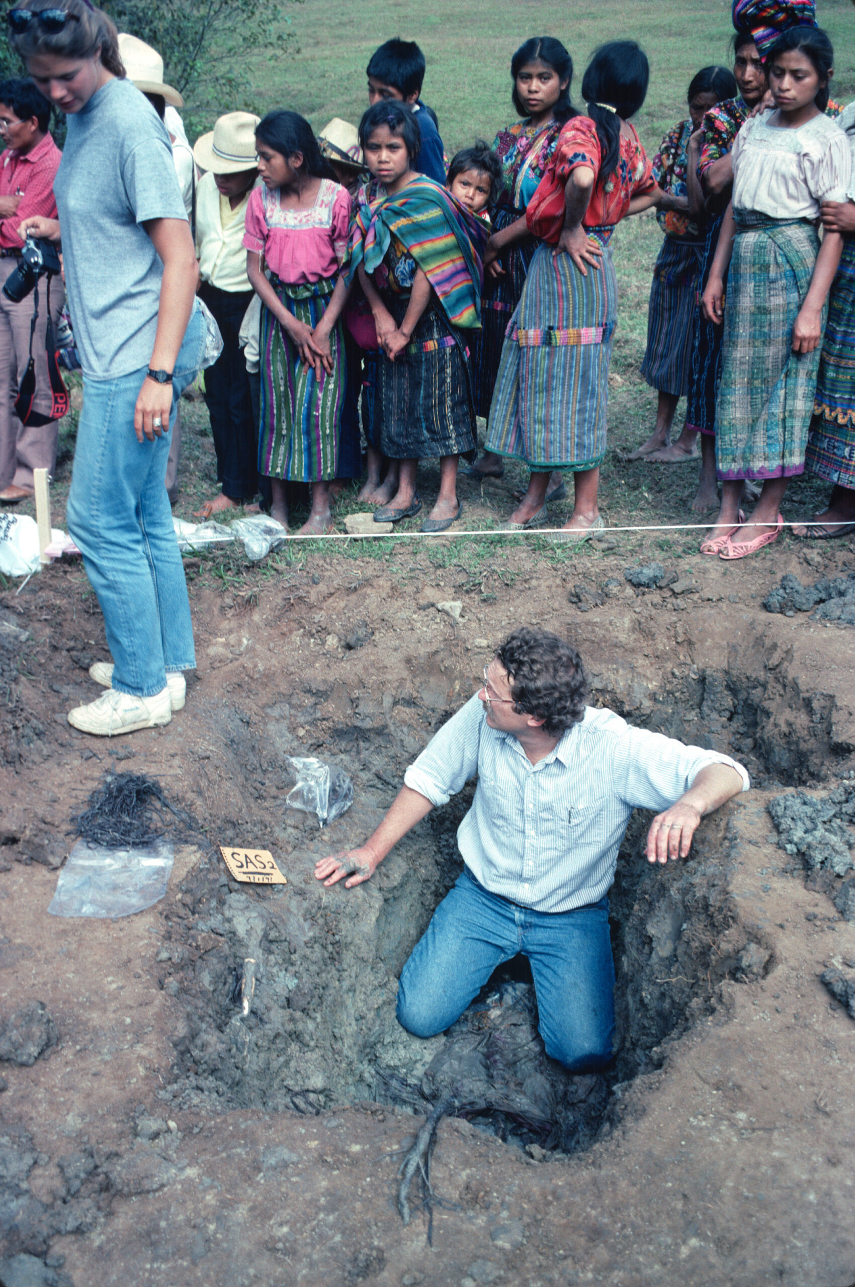 A man in a dirt hole looks up at community members watching him participate in a forensic dig.