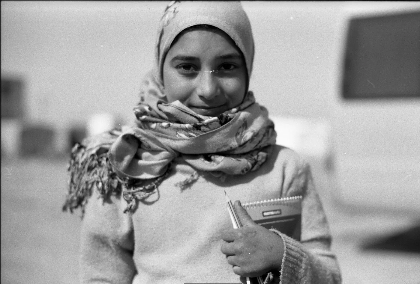 A young girl in a hijab smiles at the camera. She is holding a notebook and pencil.
