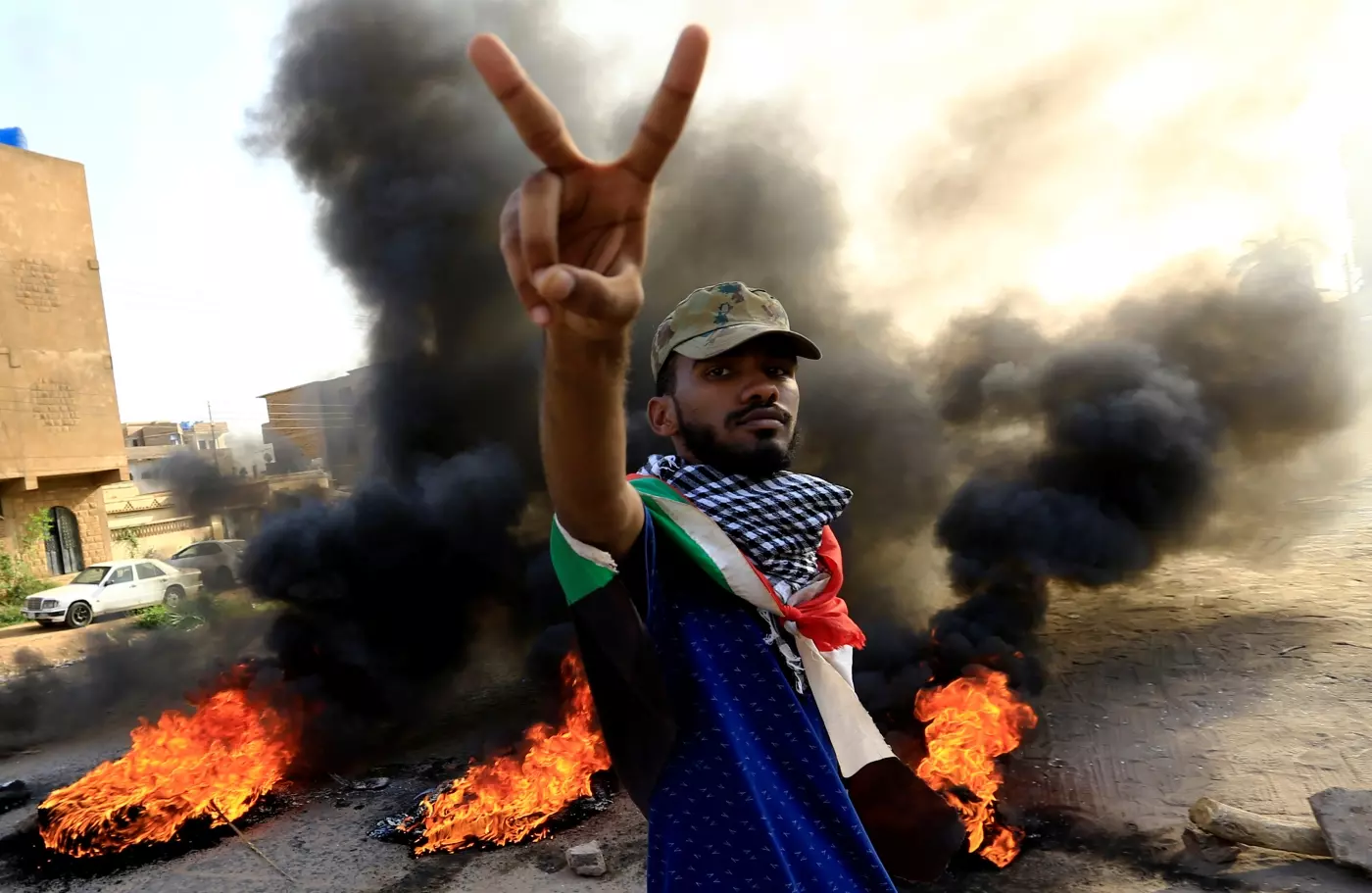 A man with the Sudan flag tied around his neck shows the camera a peace sign, while a car burns behind him.