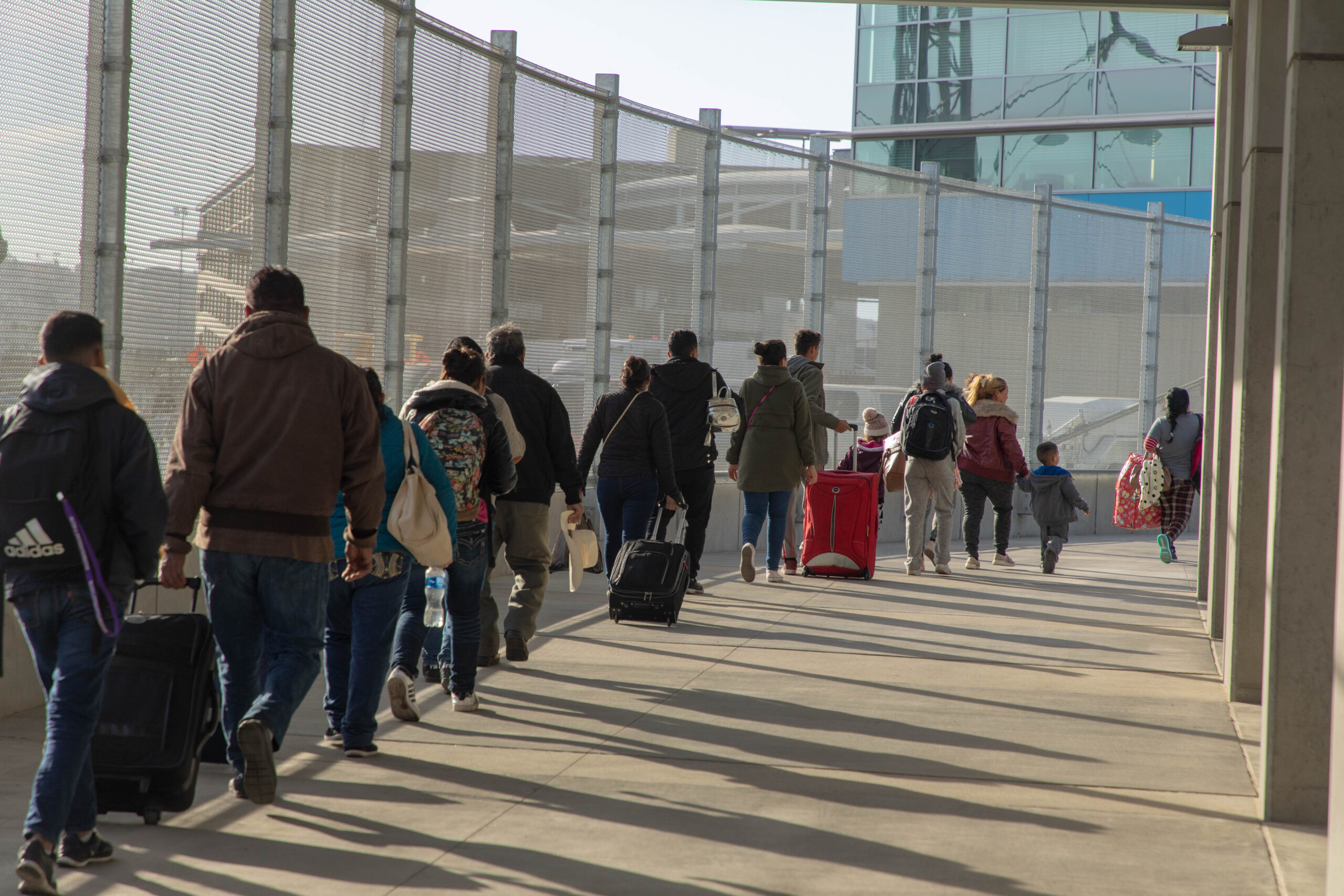 People walk in a line, some with suitcases, near a fence.