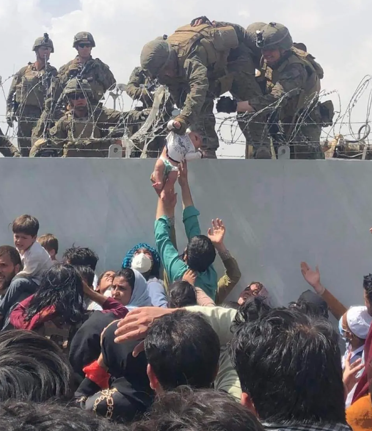 A U.S. Marine lifts an infant over a barbed wire fence during an evacuation at Hamid Karzai International Airport in Kabul. (Photo by - / Courtesy of Omar Haidiri / AFP).