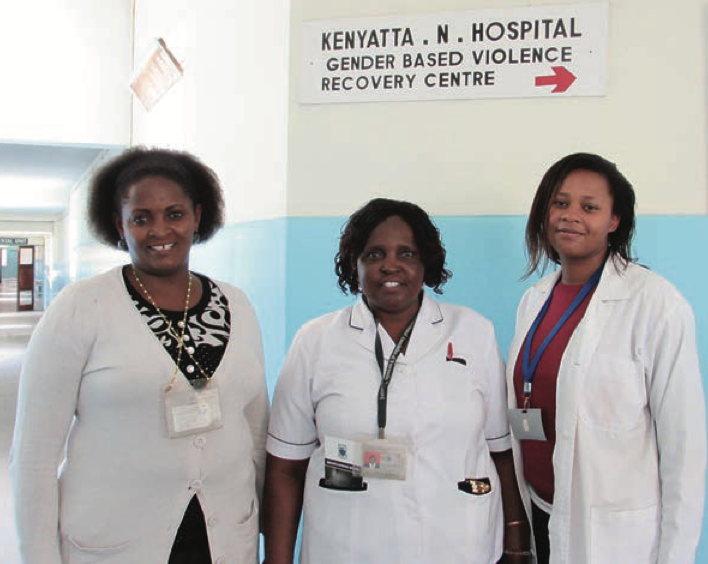 Three woman in white coats with brown hair smile at the camera. They are at a hospital.
