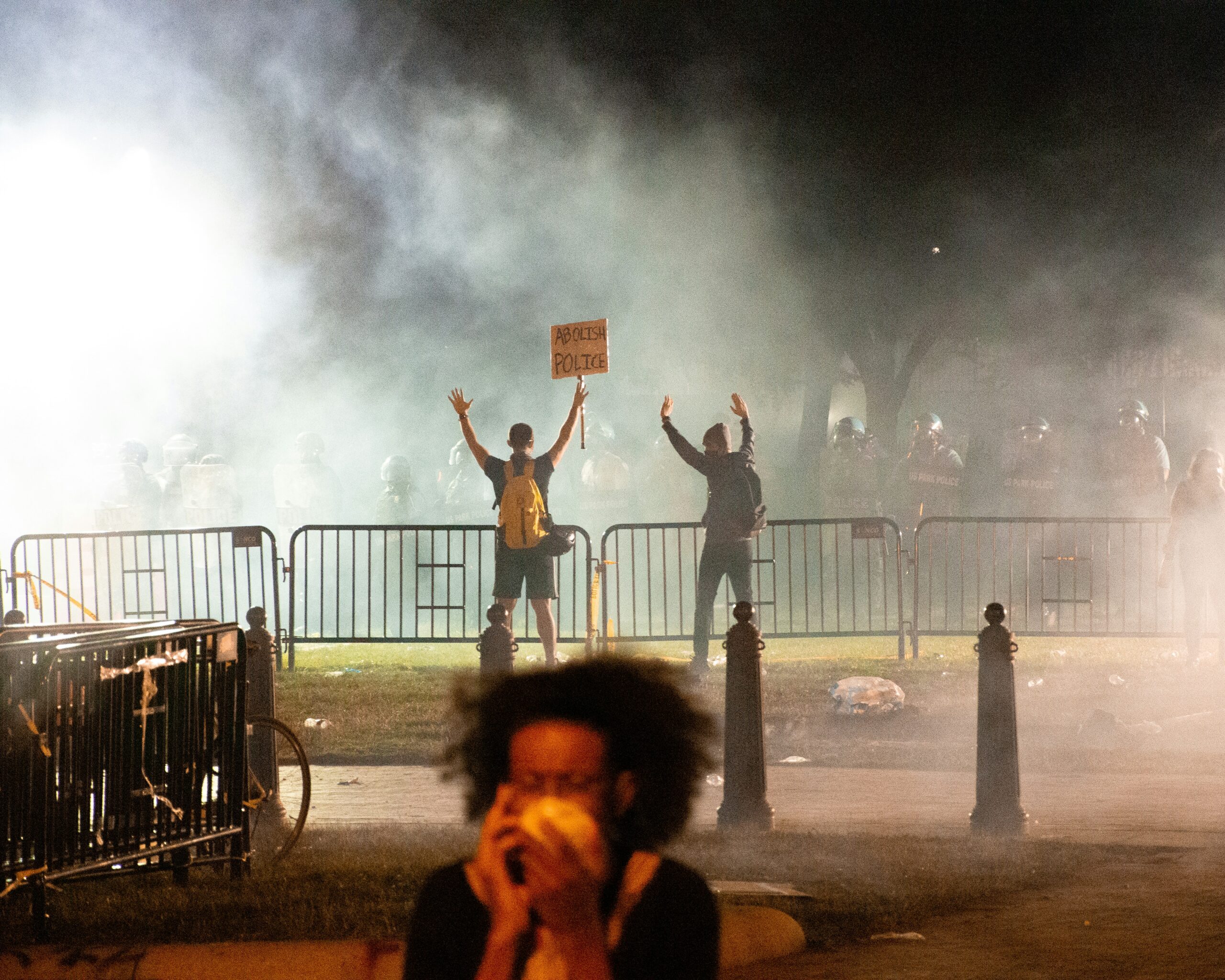 A protest with tear gas in the air. One protester is holding a mask to her face to avoid breathing in the gas. Two protesters in the background have their hands raised, one holds a sign that says 