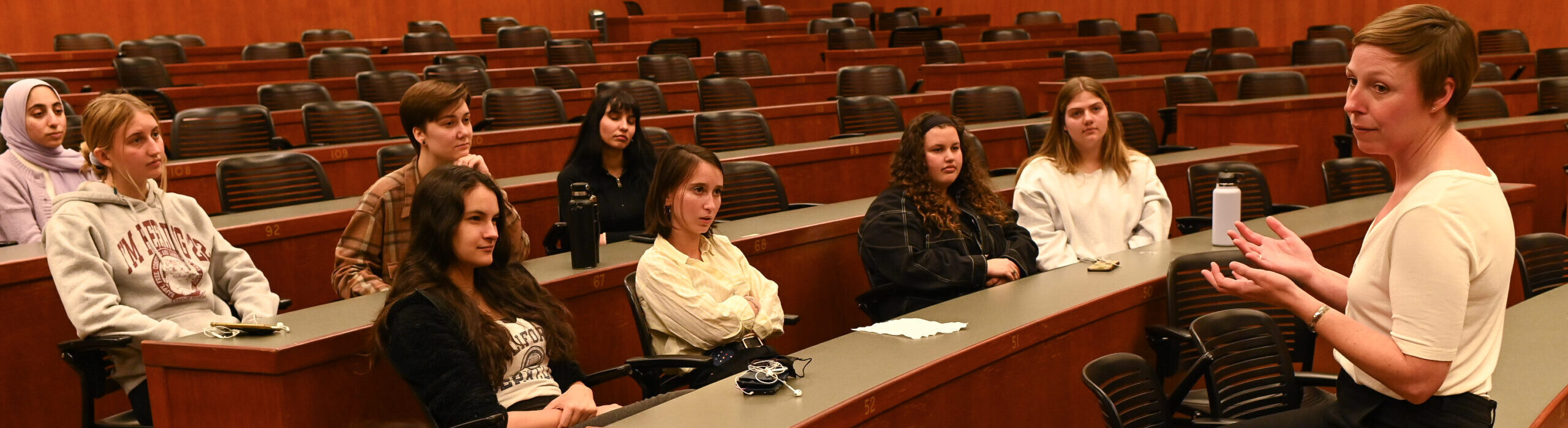 A woman speaks to a group of young adults in a large room.