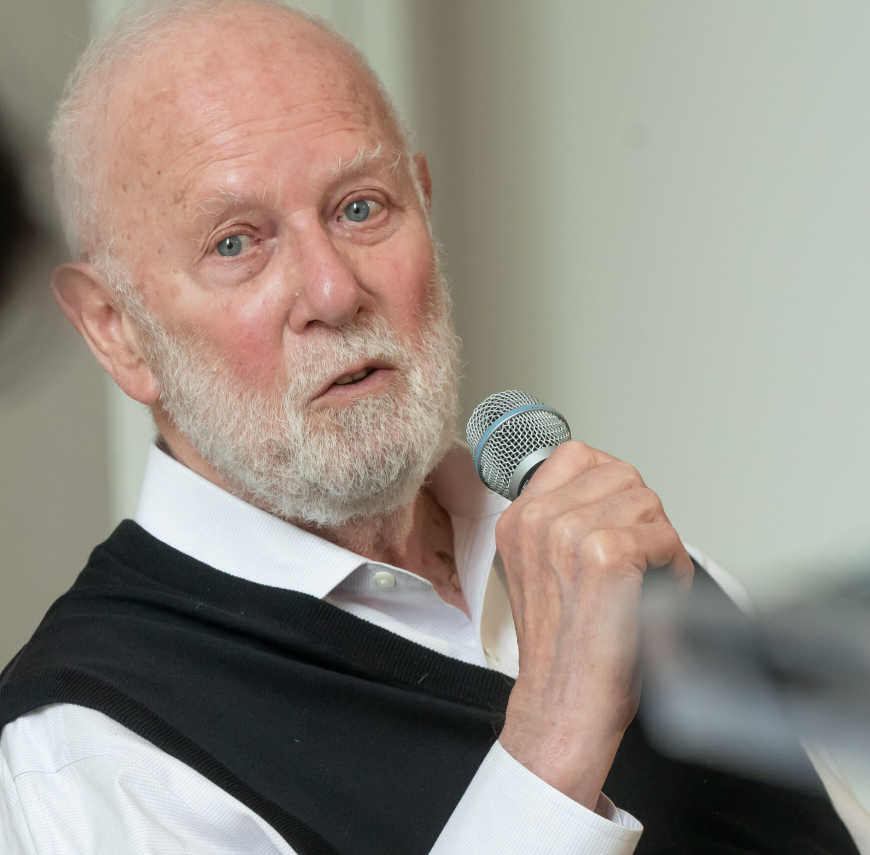 A man speaks into a microphone. He was white hair and a white beard.