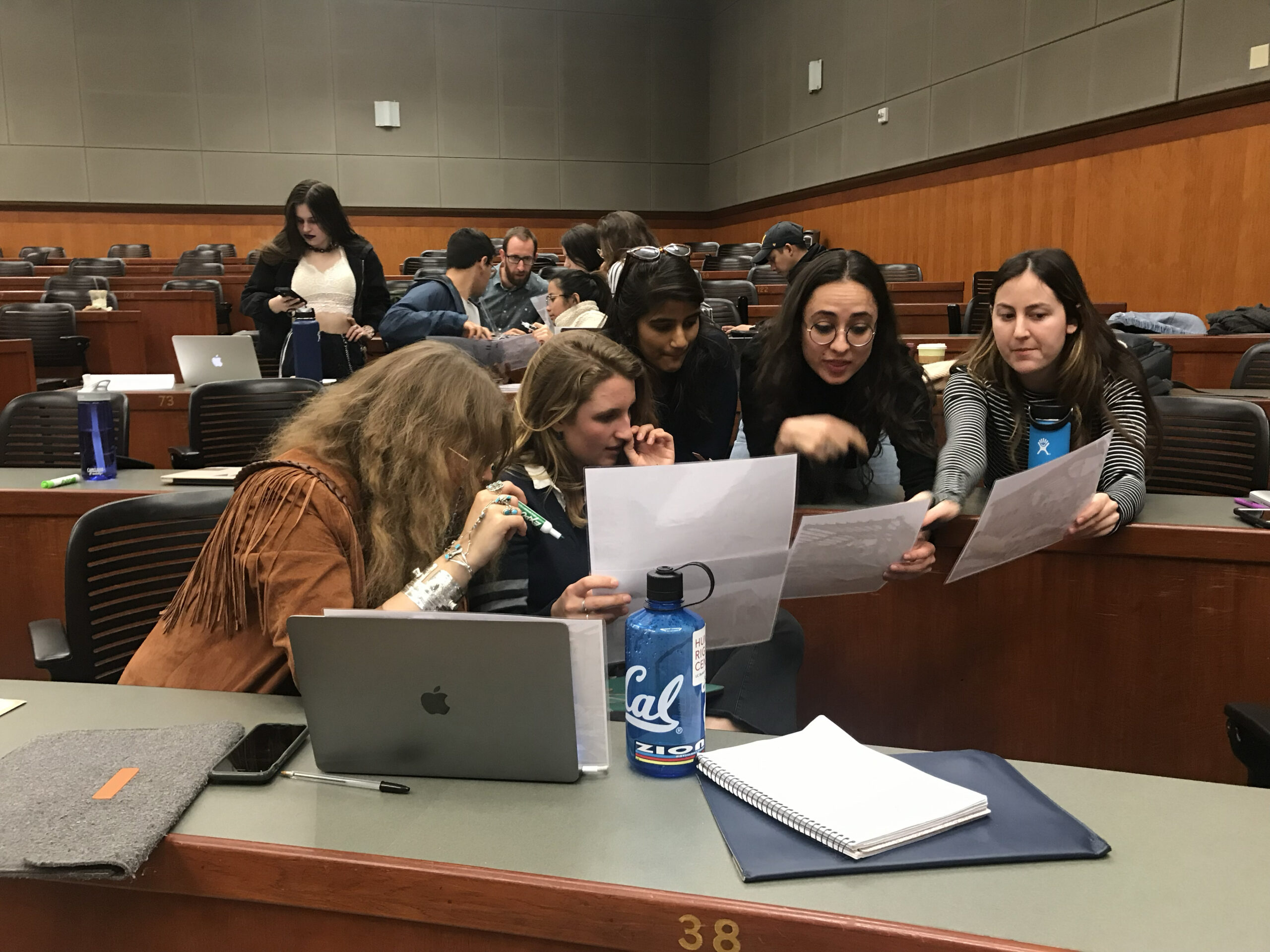 A group of people sit in an auditorium, there is a laptop and several papers.
