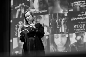 A woman speaks into a microphone and looks down at a notecard. Behind her, images of people with eyepatches are projected in a collage.