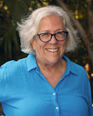 A woman smiles at the camera. She has glasses on and white hair.
