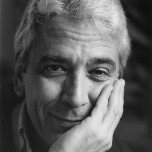 A man with white hair holds his hand to his cheek and smiles pensively at the camera.
