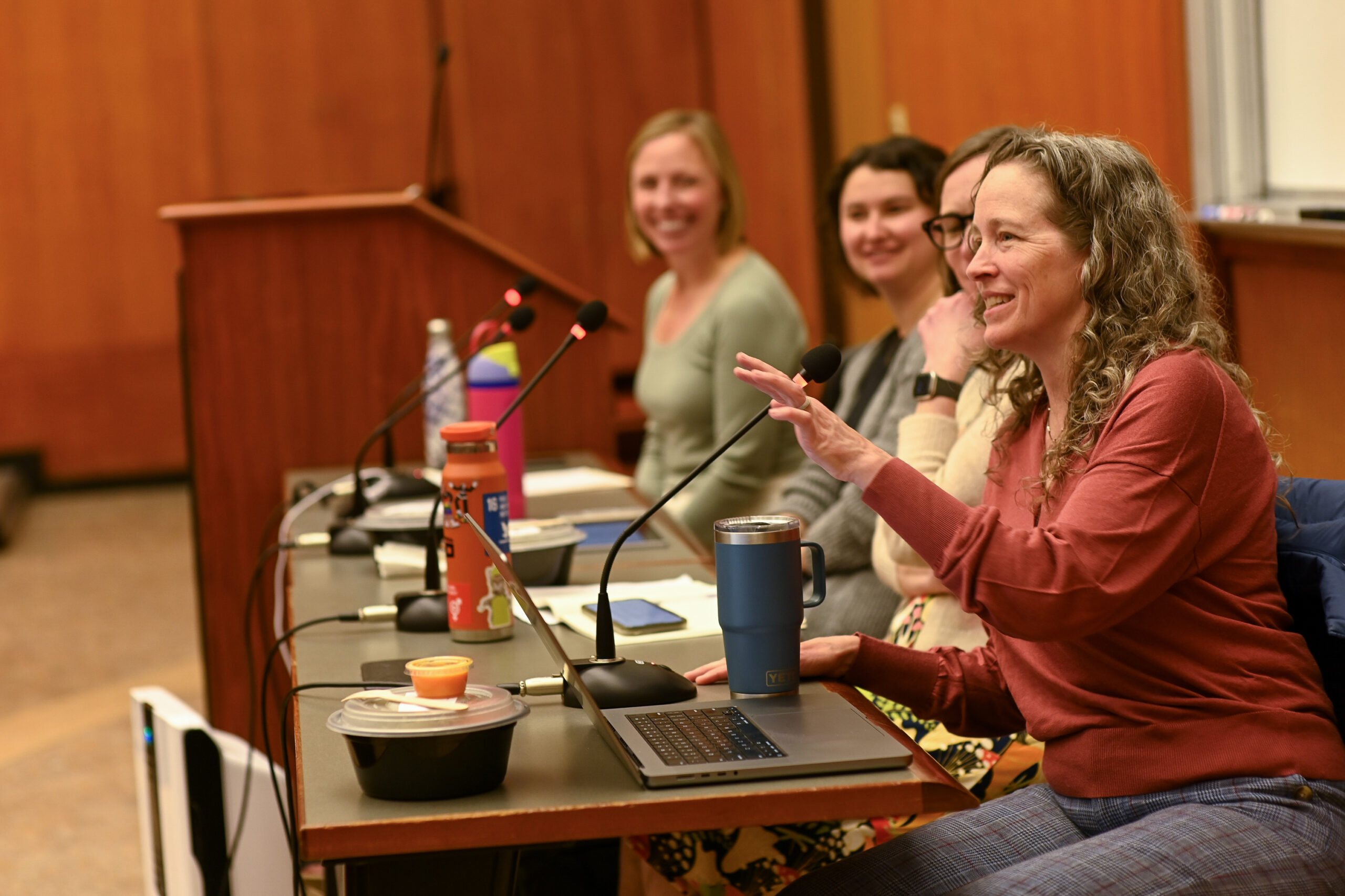 Four women sit on a panel, one of them speaks and the others smile.