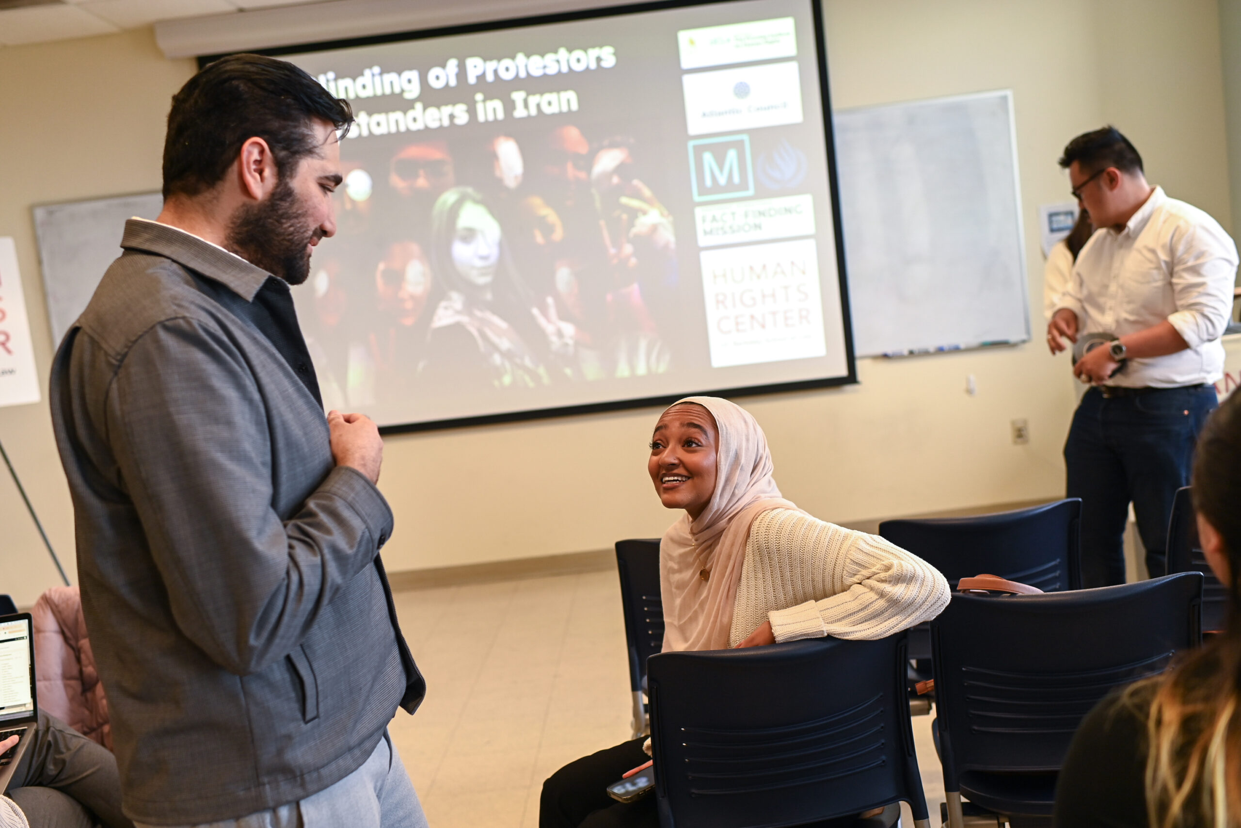 A man standing up speaks to a woman sitting down. There is a presentation behind them that reads: 