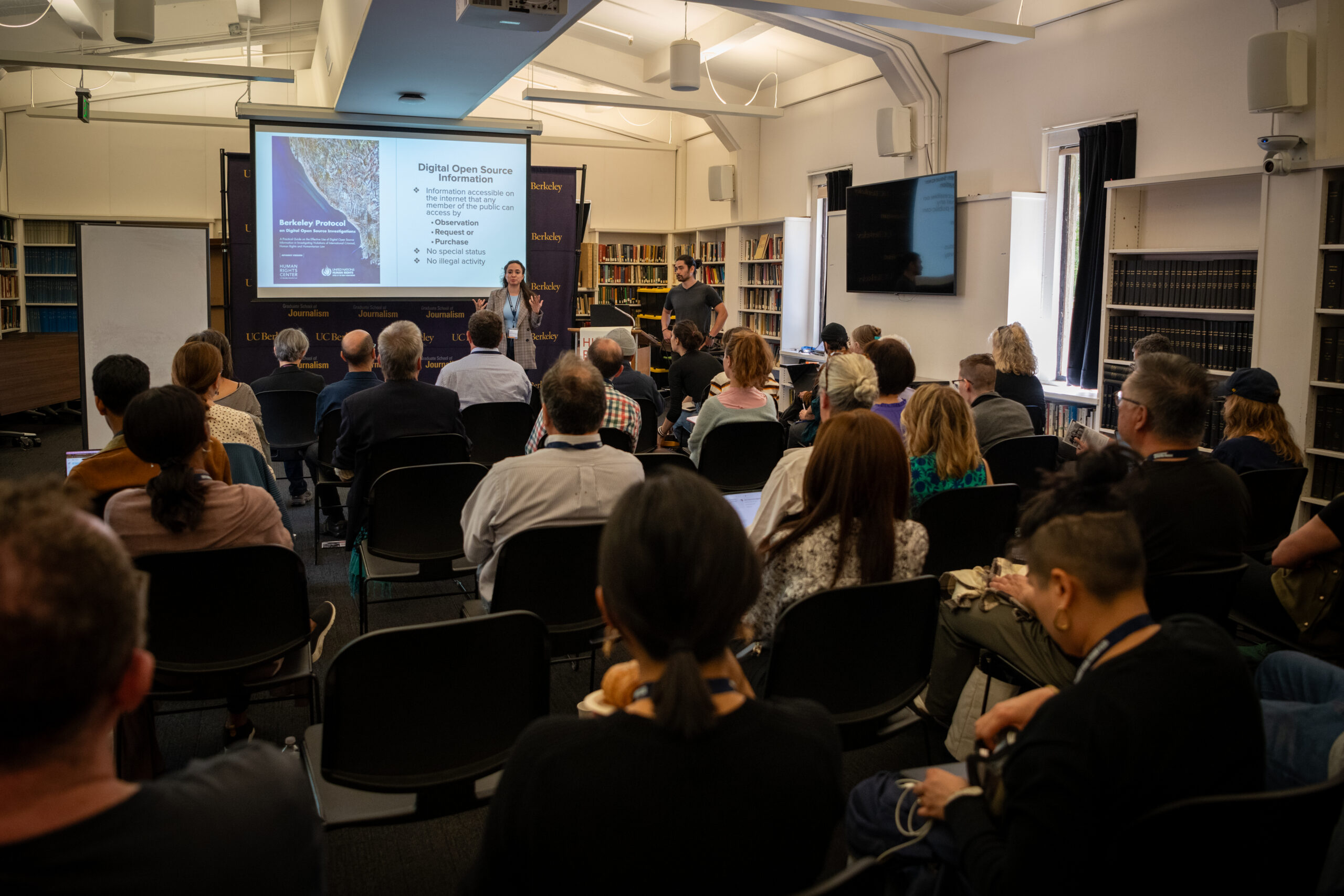 A group of people in a room look at a man and a woman presenting. There is a presentation that reads 