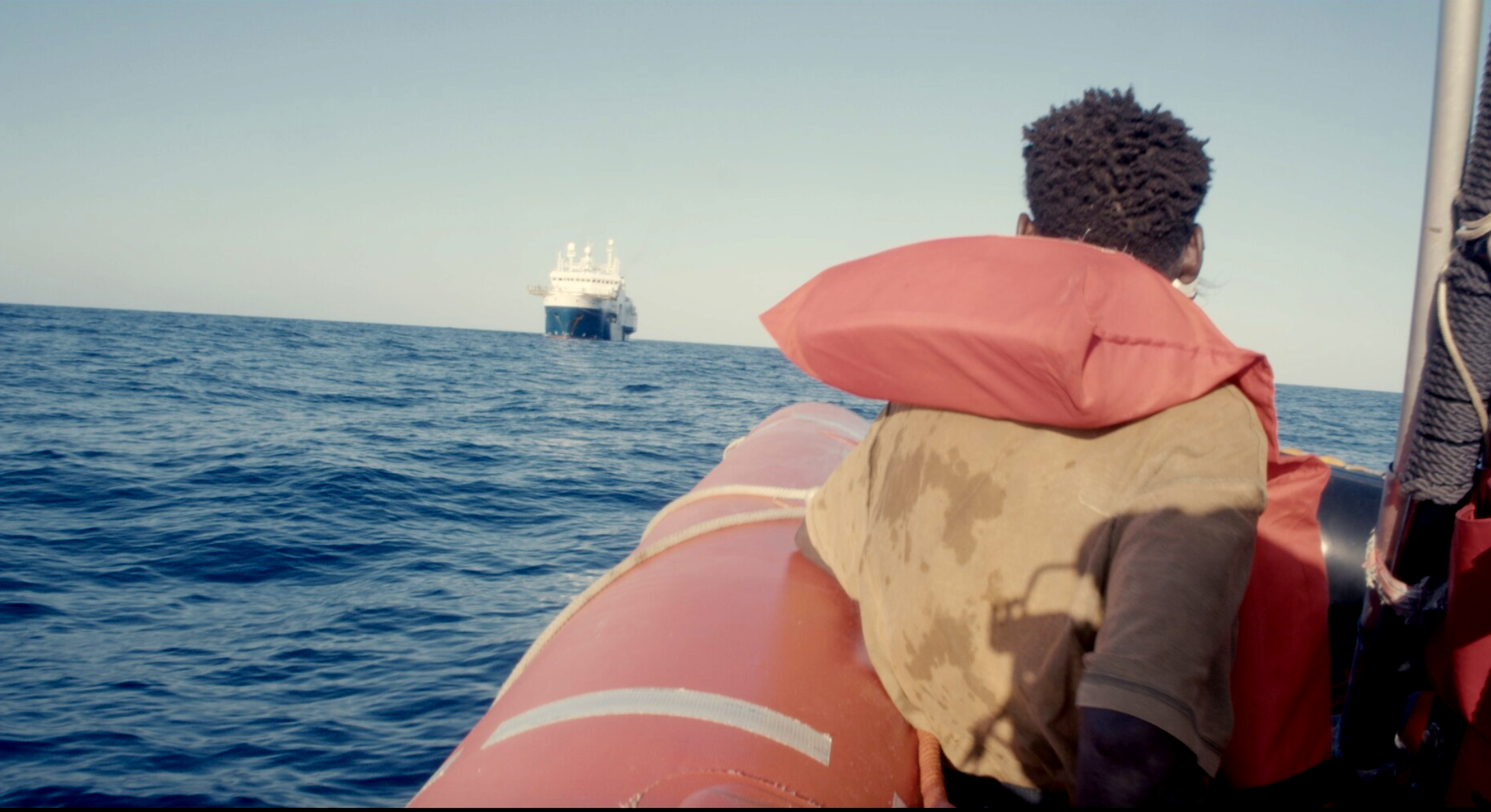 A man in a rubber boat wearing a life vest on the ocean looks to the distance at a large boat.