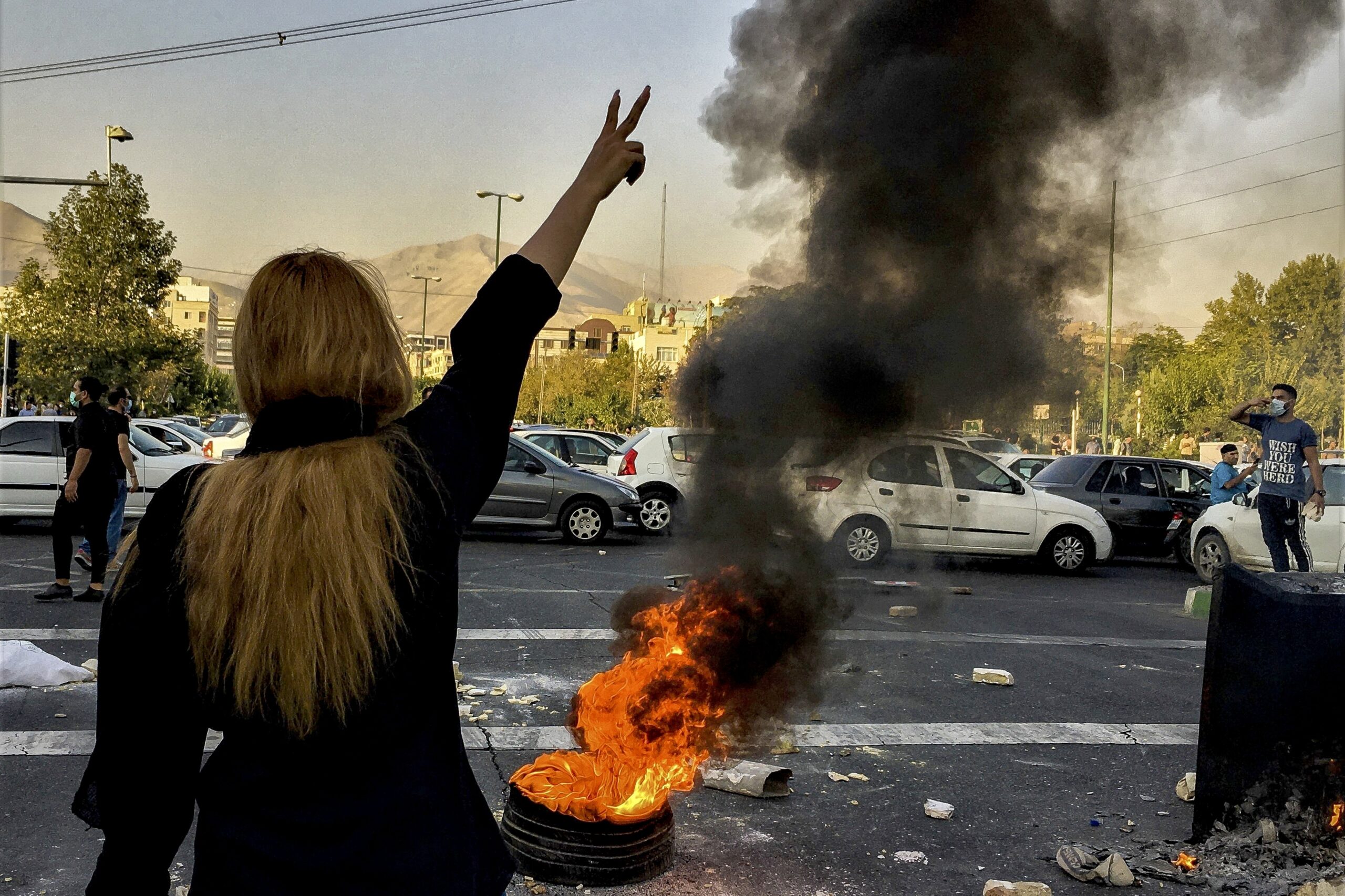 A woman holds her two fingers up in a victory symbol. In front of her, a tire burns. Her face is not visible.