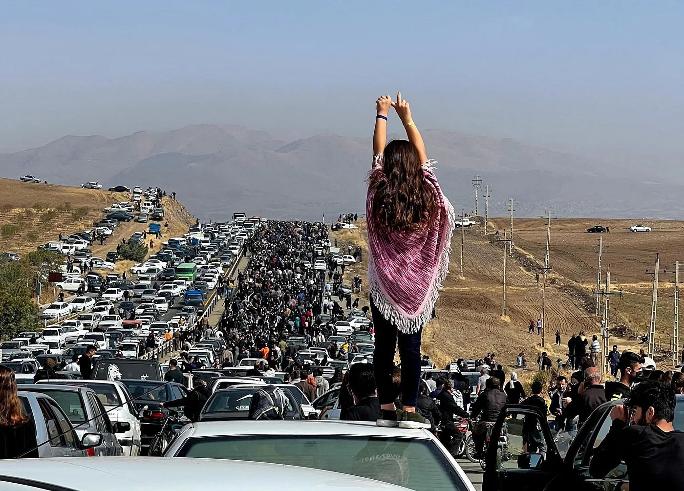 A young girl stands on a car in a sea of protesters. She holds her middle and pointer finger up, indicative of the victory sign in Iran during the Woman, Life, Freedom movement.