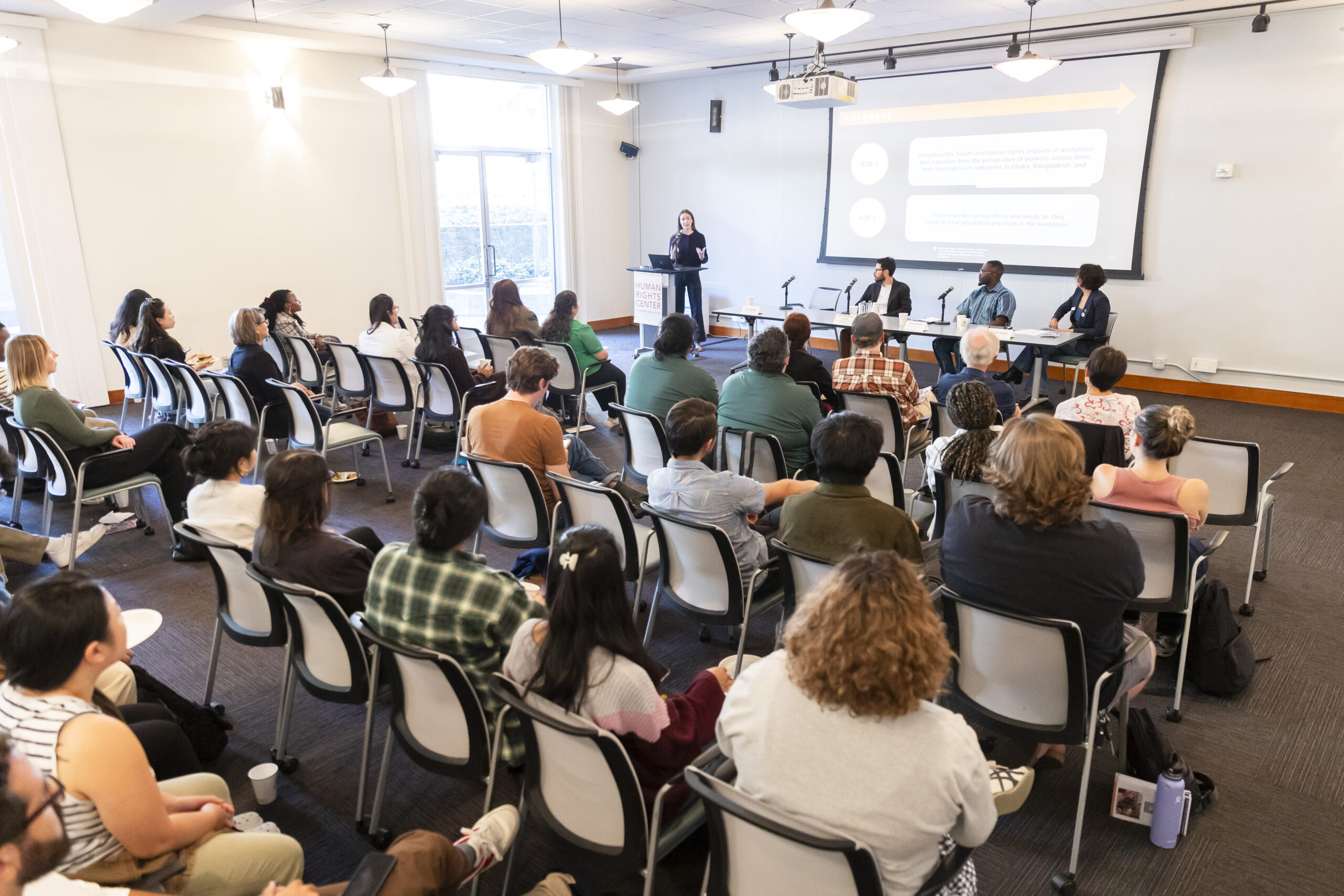 A large audience faces a presenter. The presenter is projecting a powerpoint.