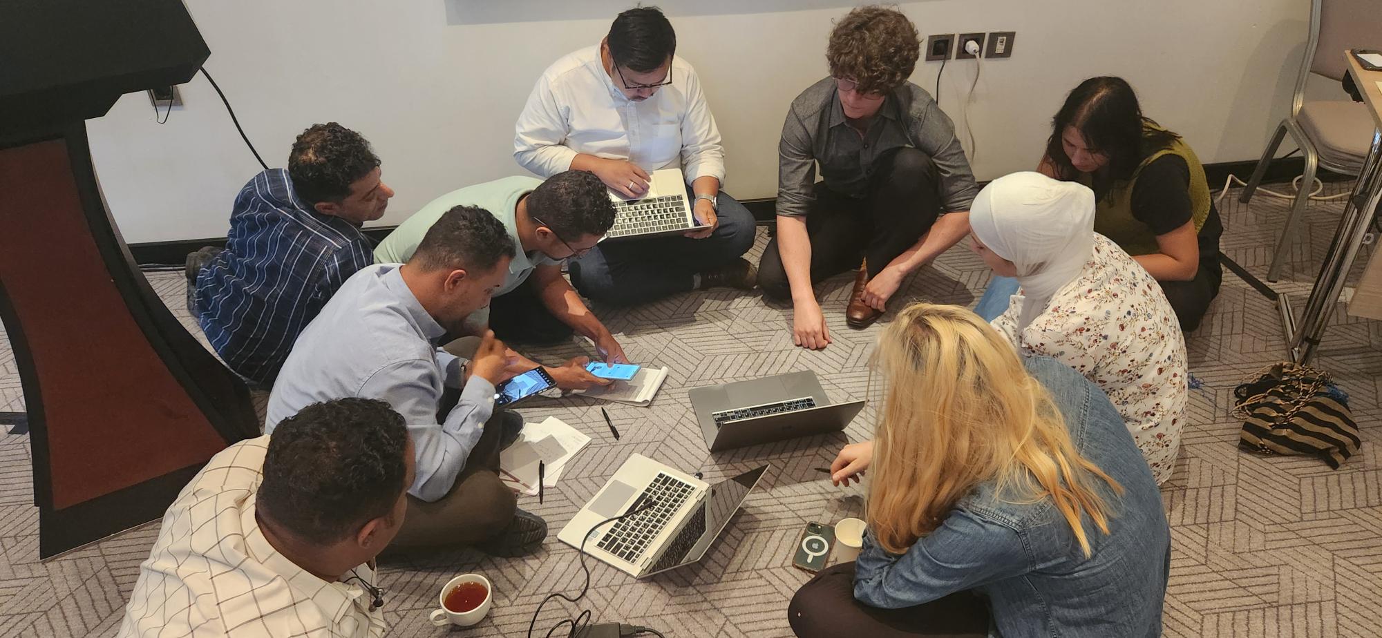 Nine people sit on the floor, their faces are shielded and they work on phones and computers. There is a mixture of women and men.