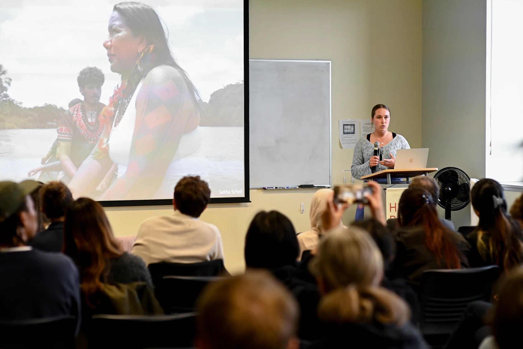 A young woman presents to a group of people from a podium. Behind her, a screen projects the image of two people in a boat.