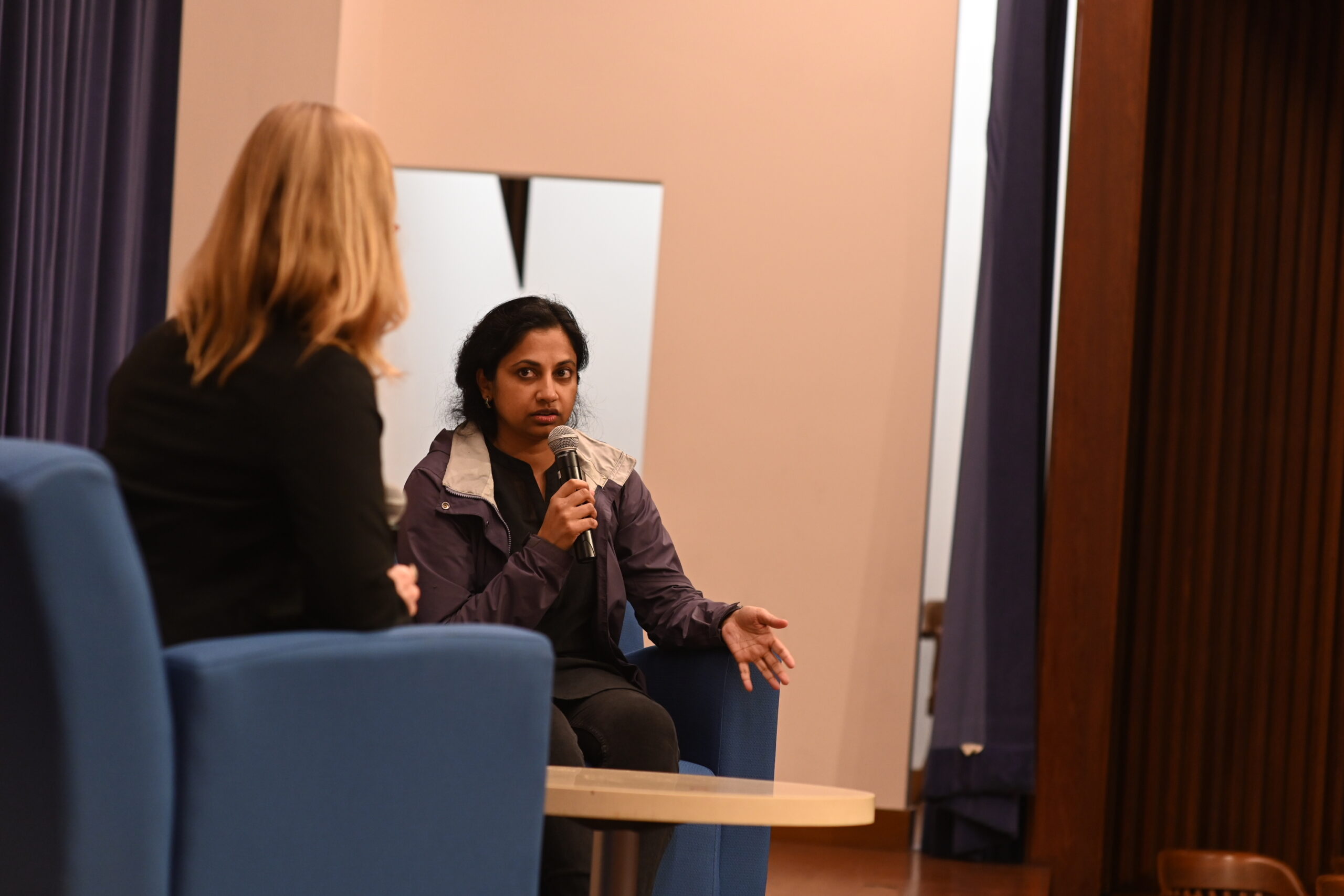 A woman speaks into a microphone on a stage, and another woman looks at her.